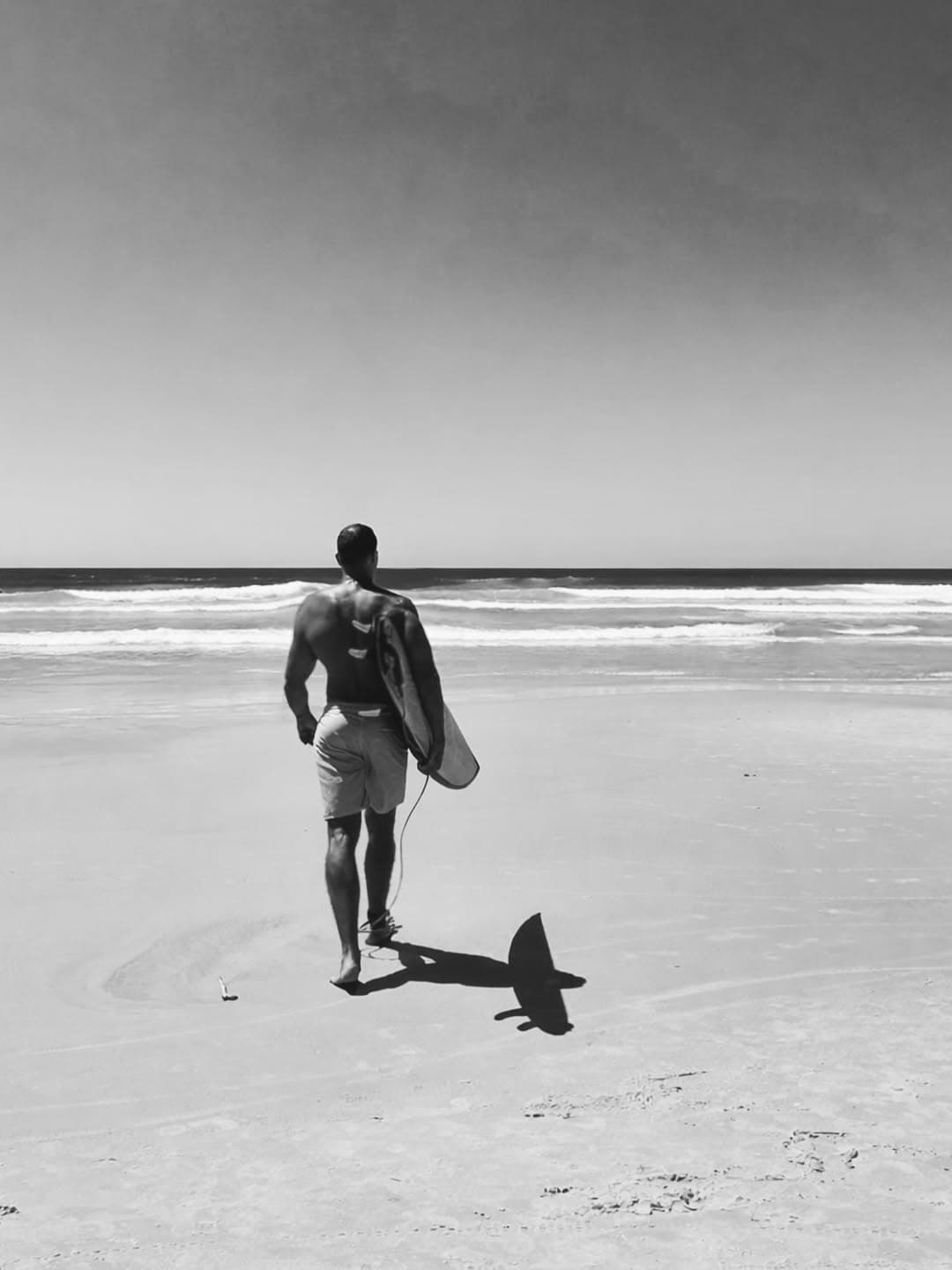 Dr. Marcelo Nunes caminhando na praia com sua prancha de surf, em preto e branco